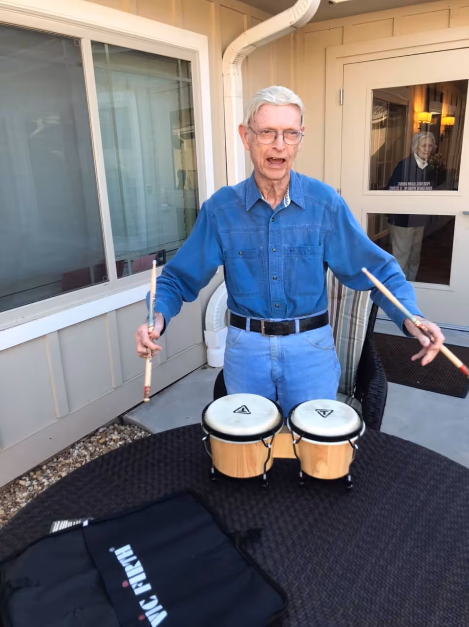 An elderly man wearing a blue shirt and jeans is standing outdoors at a round table playing a pair of bongo drums with drumsticks. Behind him is a building with a window and a door, and a reflection of another person is visible in the door's glass.