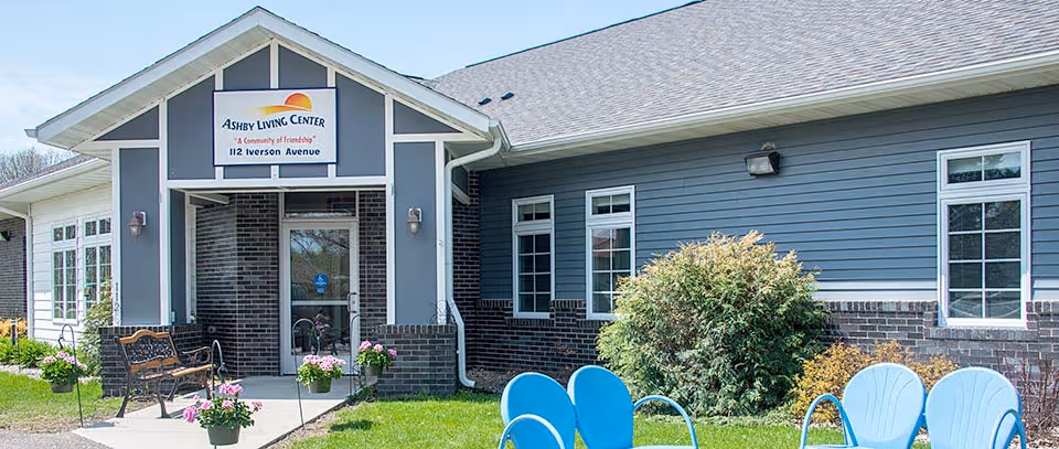 Exterior view of Ashby Living Center building with a sign above the entrance reading 'Ashby Living Center, A Community of Friendship, 112 Iverson Avenue'. The building has blue siding with brick accents and white trim. There are benches and flower pots near the entrance, and blue outdoor chairs on the grass in front of the building.