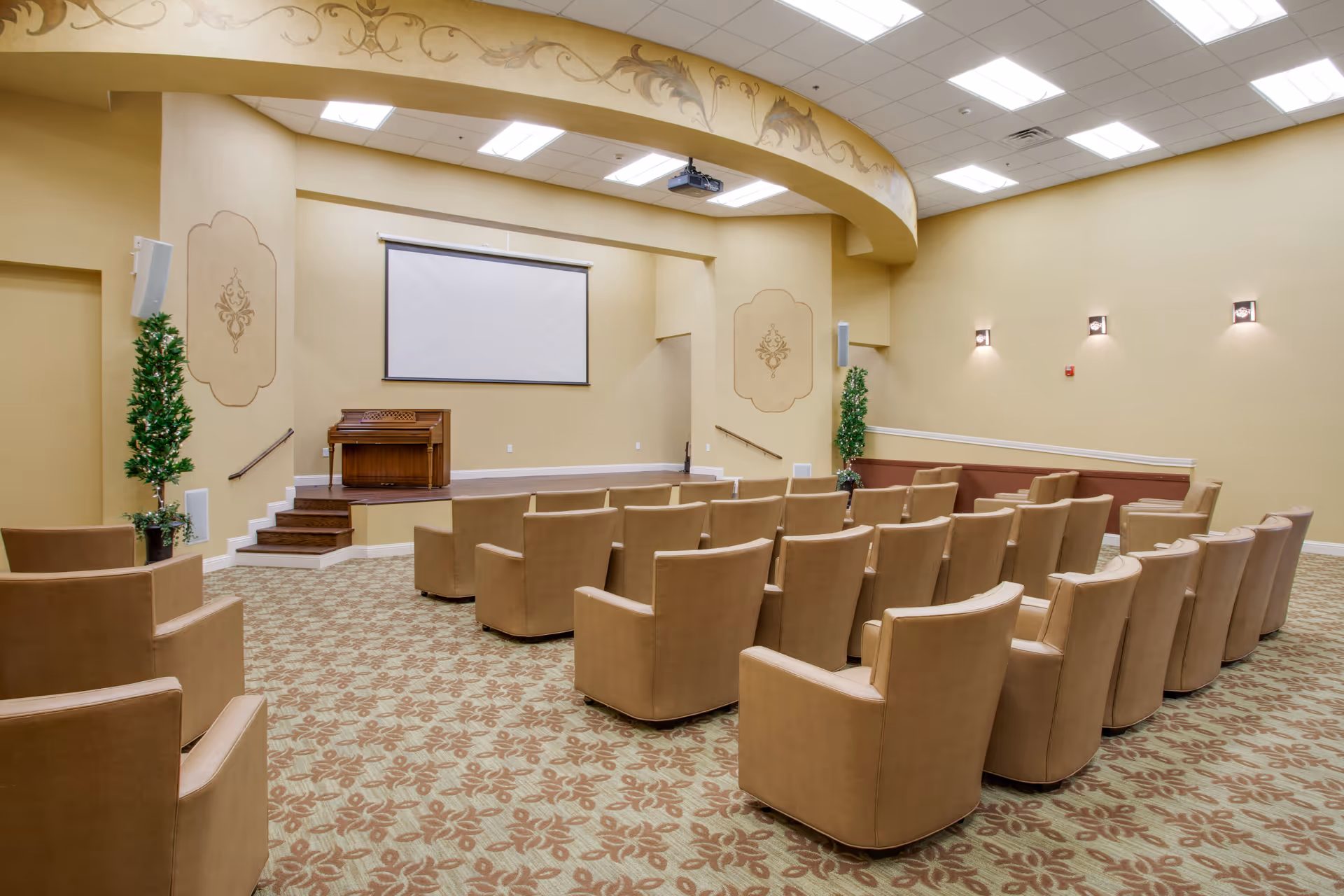 A small auditorium or presentation room with rows of beige upholstered chairs facing a stage. The stage has a wooden organ and a large white projection screen mounted on the wall. The room features beige walls with decorative wall sconces and two potted plants on either side of the stage. The ceiling has recessed lighting and a projector mounted above the stage.