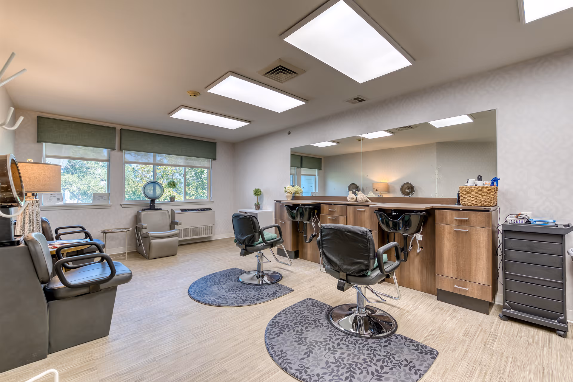 A bright and clean hair salon area inside a senior living facility with two black salon chairs in front of a large mirror and two hair washing sinks. There are additional seating chairs along the wall near windows with green blinds, a lamp, and a small side table. The room has light-colored flooring and walls with overhead rectangular lights.