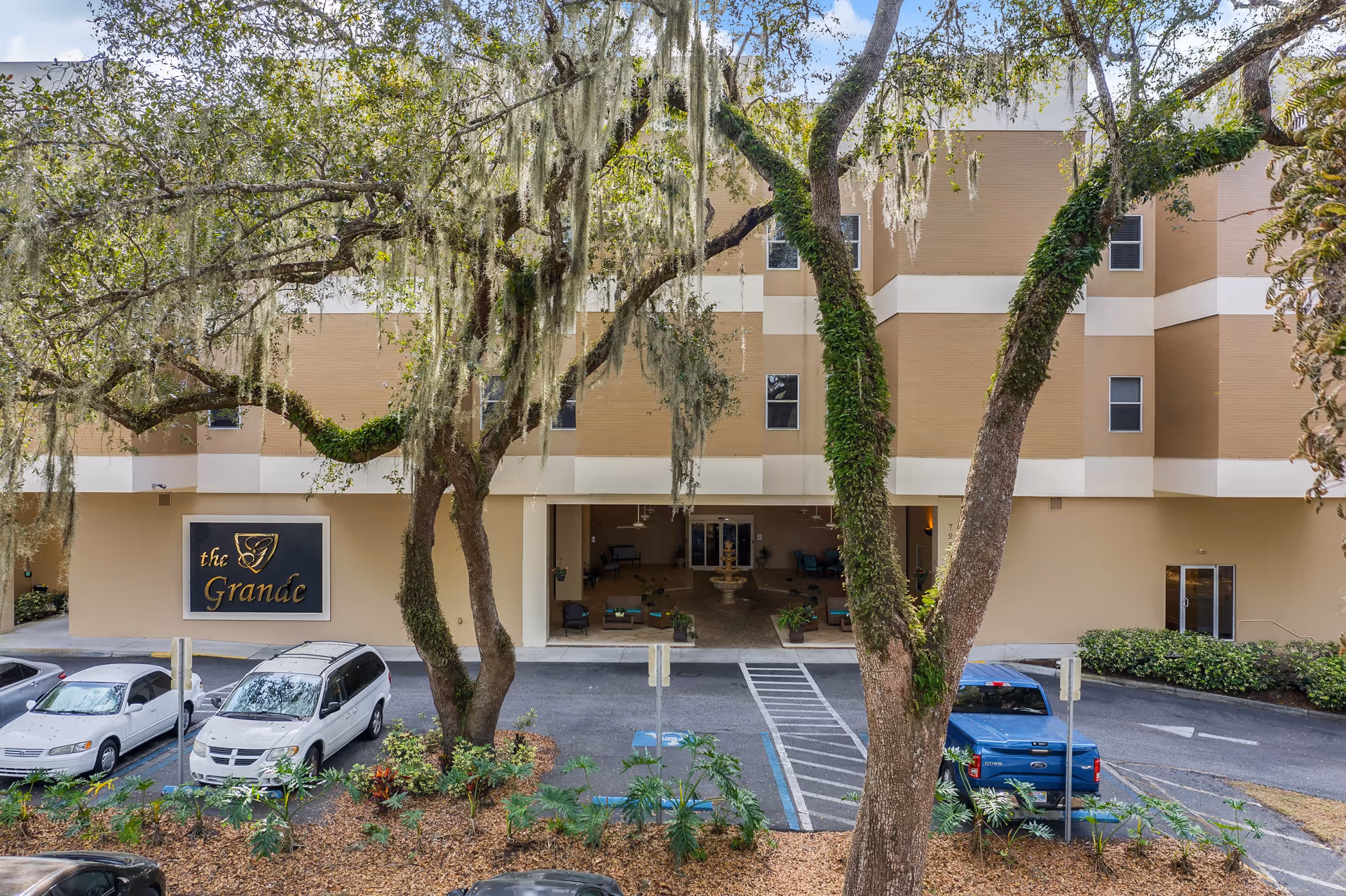 Exterior view of The Grande senior living facility showing the building entrance with a covered drop-off area, several parked cars, and large trees with hanging moss in the foreground.