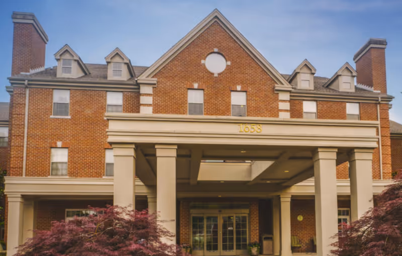 Front entrance of a three-story brick assisted living building with columns and the address number 1658 above the portico.
