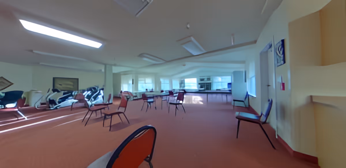 A large room with multiple chairs spaced apart on a carpeted floor, large windows letting in natural light, and a kitchen area with appliances in the background.