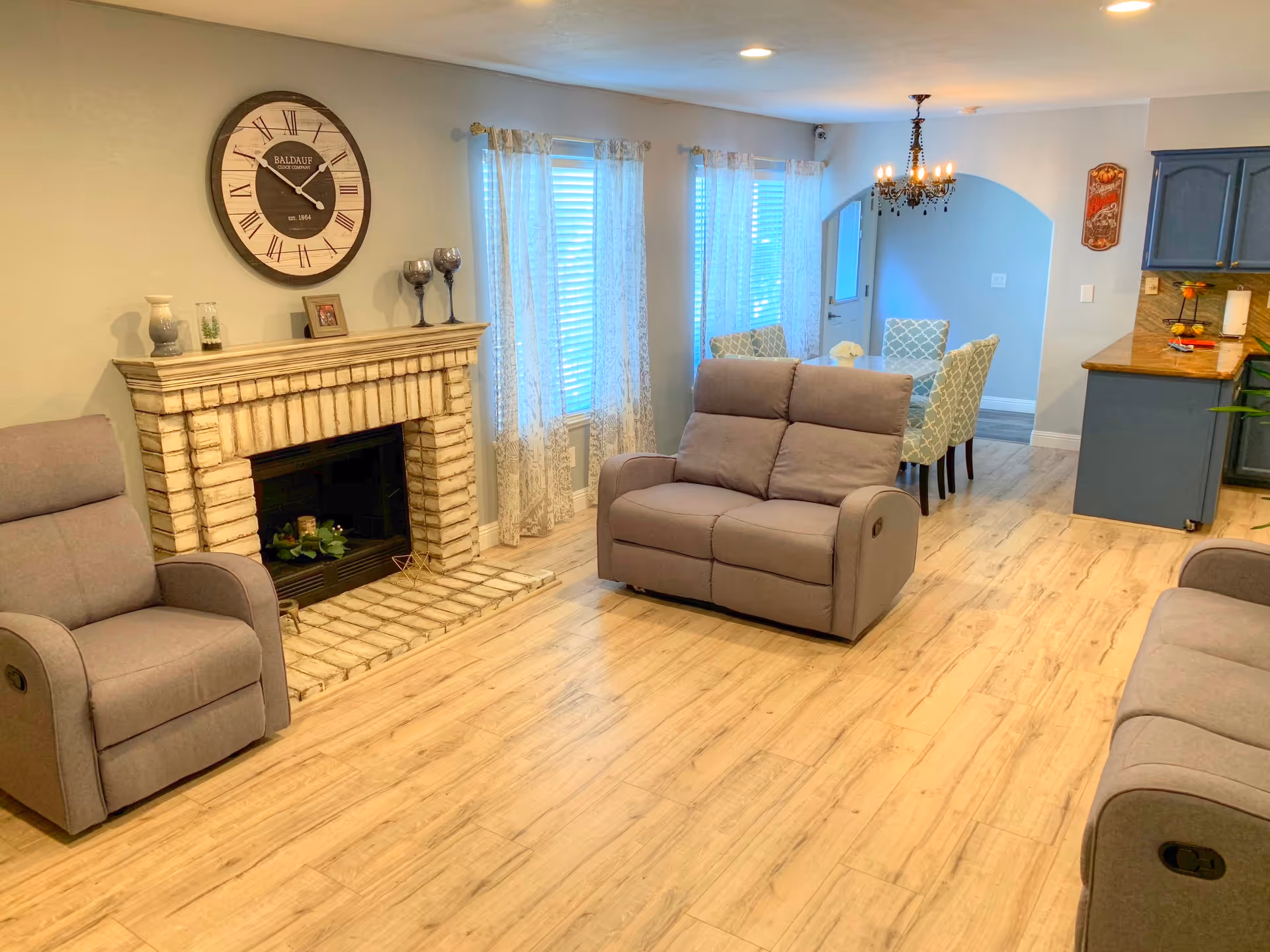 A cozy living room area with light wood flooring, a beige brick fireplace with decorative items on the mantel, a large round wall clock above the fireplace, and three gray recliner chairs. In the background, there is a dining area with a table and four patterned chairs, a chandelier, and a kitchen with blue cabinets and a wooden countertop.
