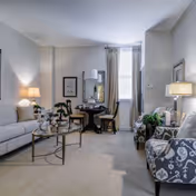 Bright living room with a sofa, patterned armchair, glass coffee table and a small dining table by a window.