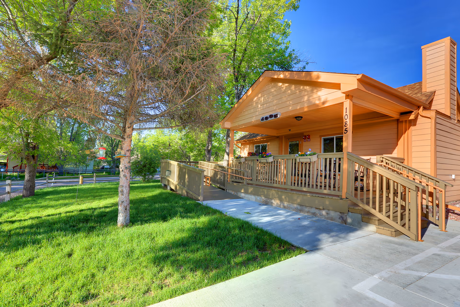 Exterior view of a single-story building with a wooden ramp and stairs leading to a covered porch. The building is painted in a warm beige color and has the number 1085 displayed on the front. There is green grass and trees surrounding the building under a clear blue sky.