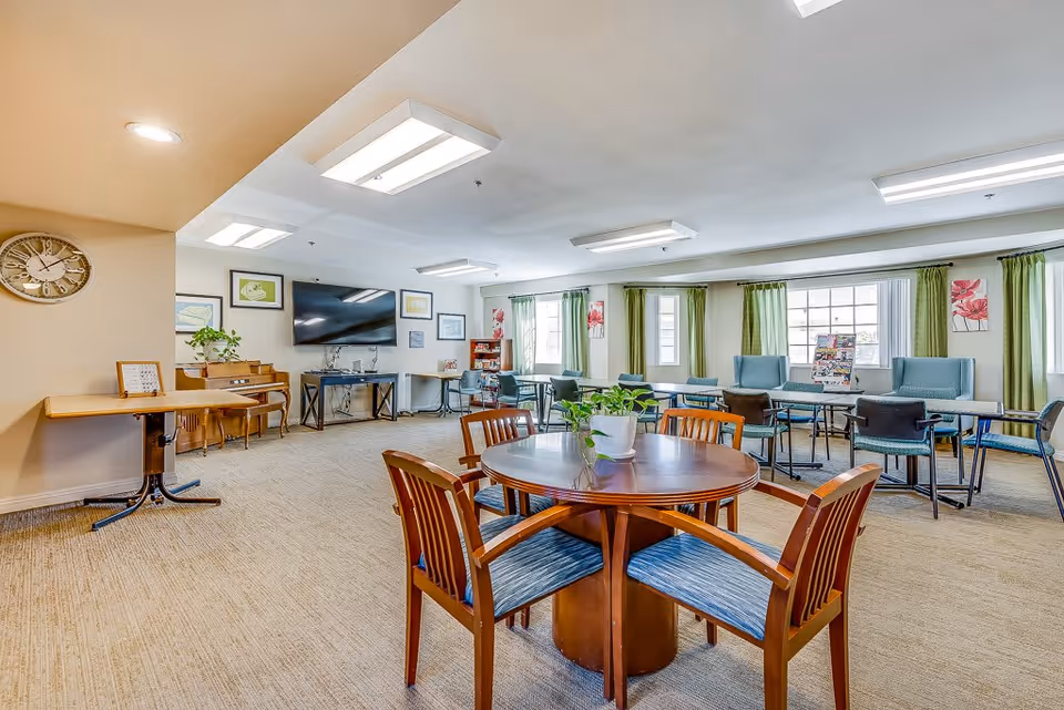 A spacious and well-lit common area in a senior living facility featuring a round wooden table with four chairs in the foreground, a piano against the wall, a large flat-screen TV mounted on the wall, several framed pictures, and multiple seating arrangements with tables and chairs near large windows with green curtains.