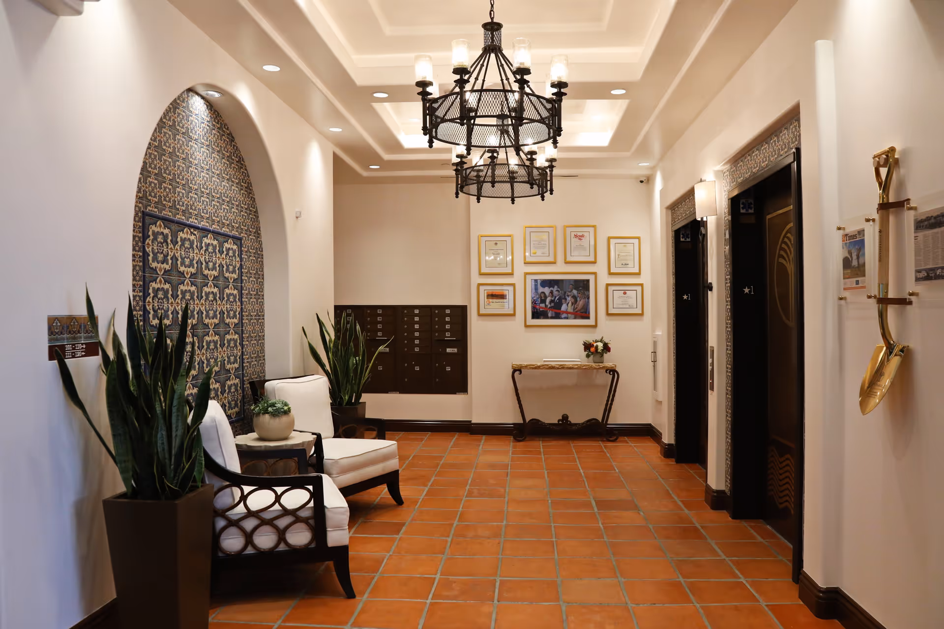 A well-lit hallway with terracotta tile flooring, two white cushioned chairs with dark wooden frames, potted plants, decorative tiled wall niche, two elevators with ornate frames, a chandelier with multiple lights, framed certificates and a photo on the far wall, and a golden ceremonial shovel mounted on the right wall.