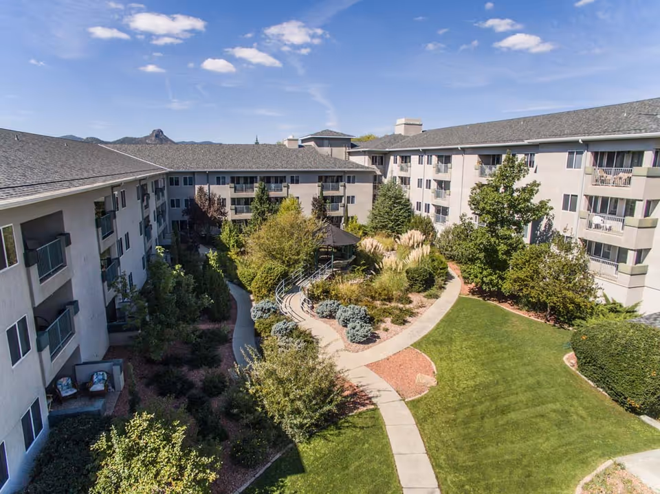 Aerial view of a senior living facility courtyard with well-maintained green lawns, walking paths, trees, shrubs, and a gazebo surrounded by a multi-story building under a blue sky with scattered clouds.