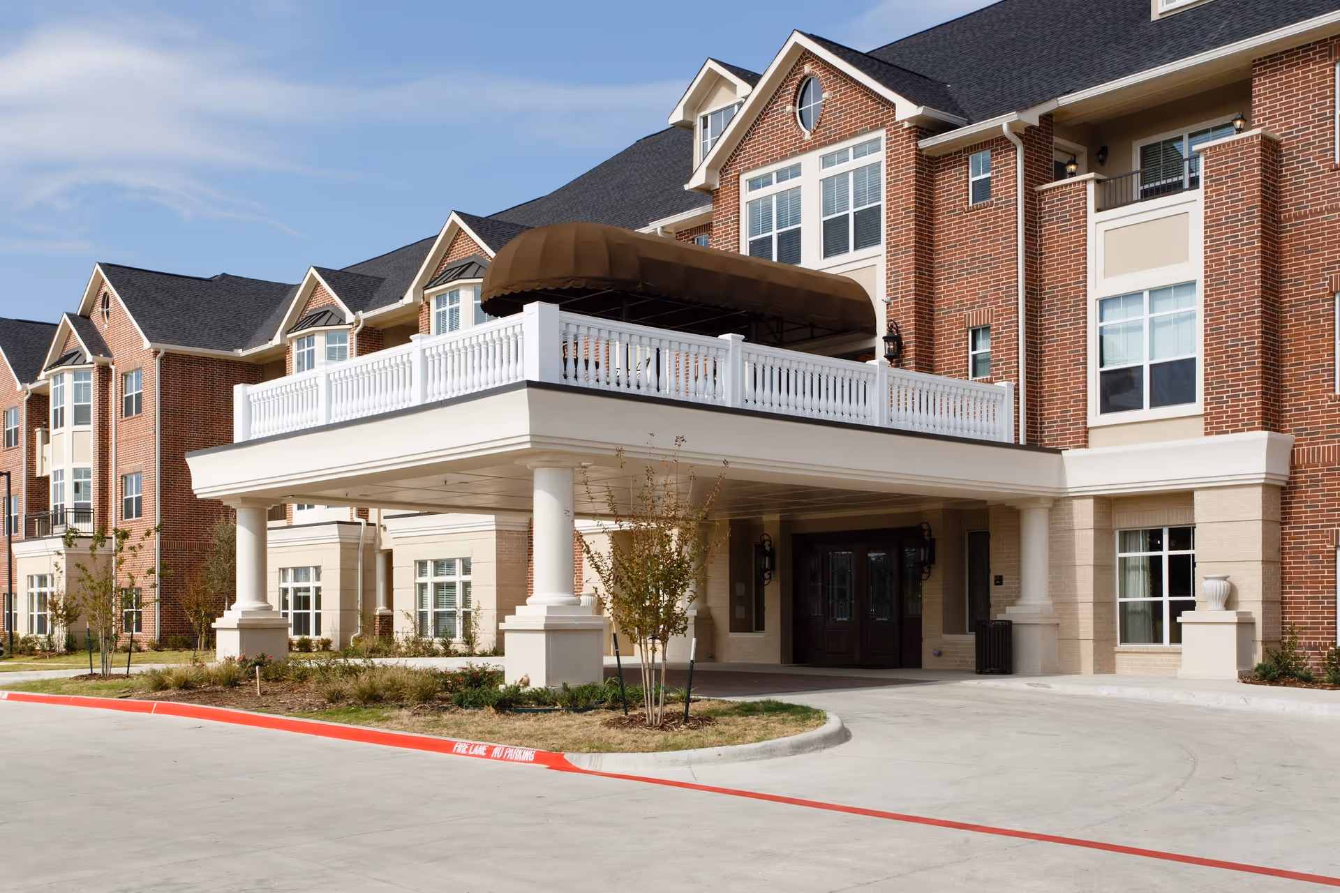 Exterior front view of a large senior living facility building with brick and beige facade, multiple windows, a covered entrance with white columns, and a balcony with a brown awning.