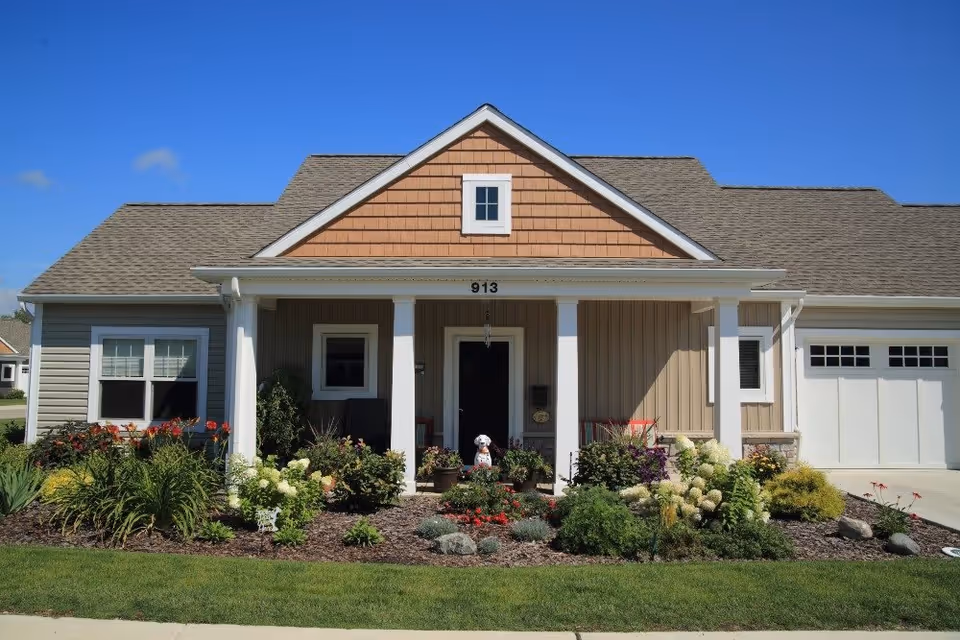 Front exterior of a single-story house with a covered porch, manicured flower beds, and an attached garage under a clear blue sky.