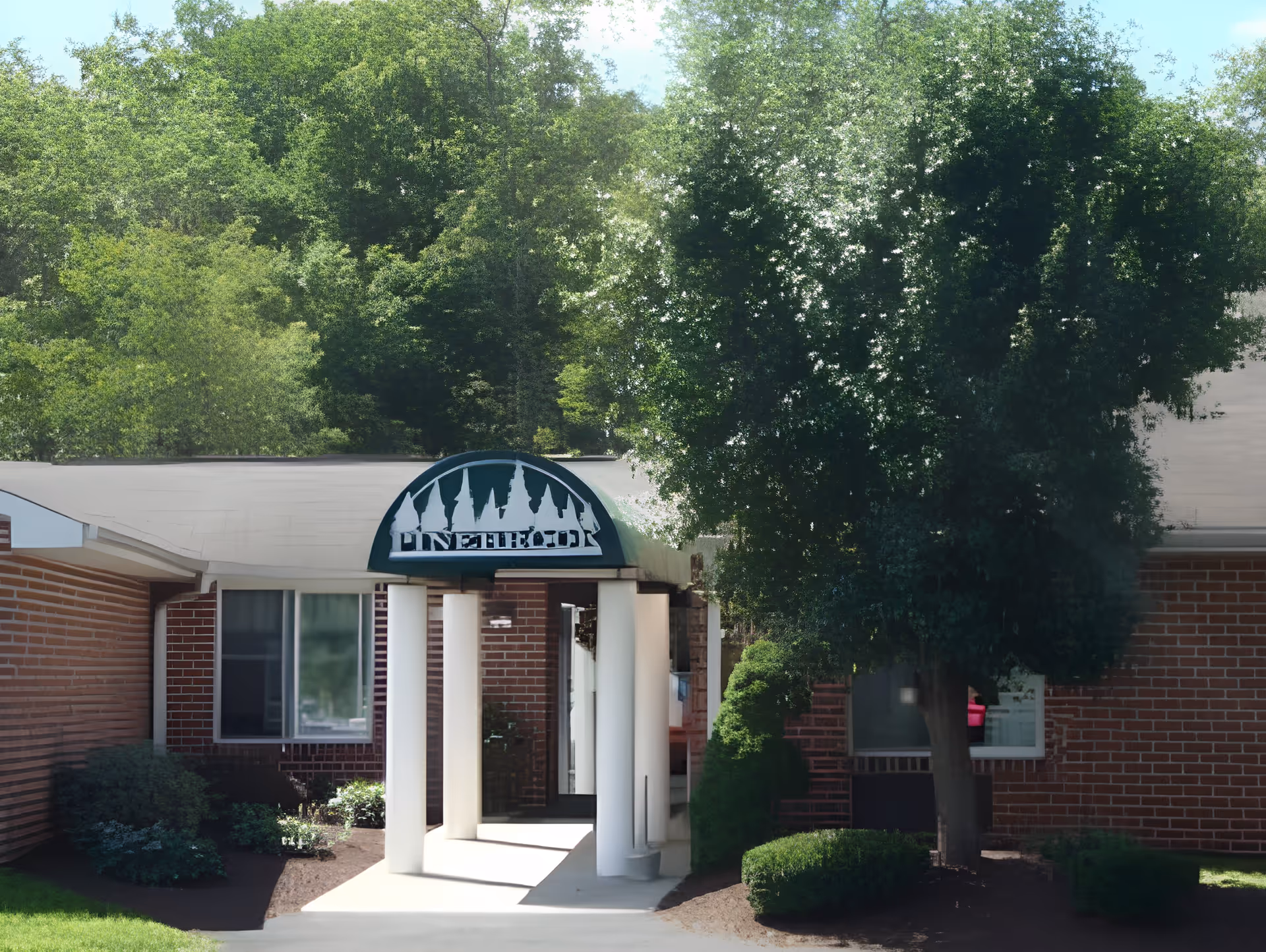 Entrance of Renaissance Home Pinebrook facility with a covered walkway supported by white columns, surrounded by green bushes and trees, and a brick building exterior.