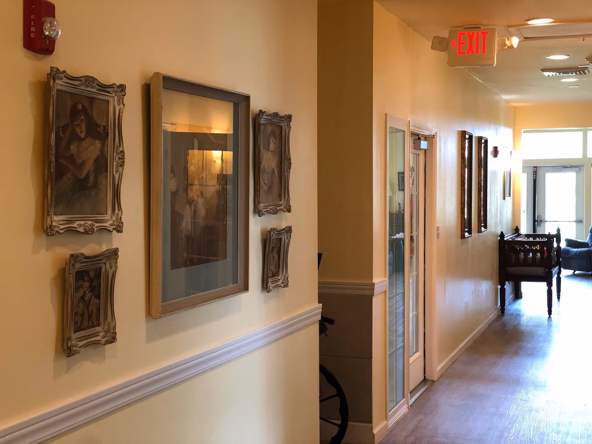 A well-lit hallway in a senior living facility with framed vintage-style paintings on the left wall and a fire alarm above them. The hallway has light yellow walls with white trim and wooden flooring. At the end of the hallway, there is a door with glass panels and an exit sign above it. Some furniture is visible near the end of the hallway.