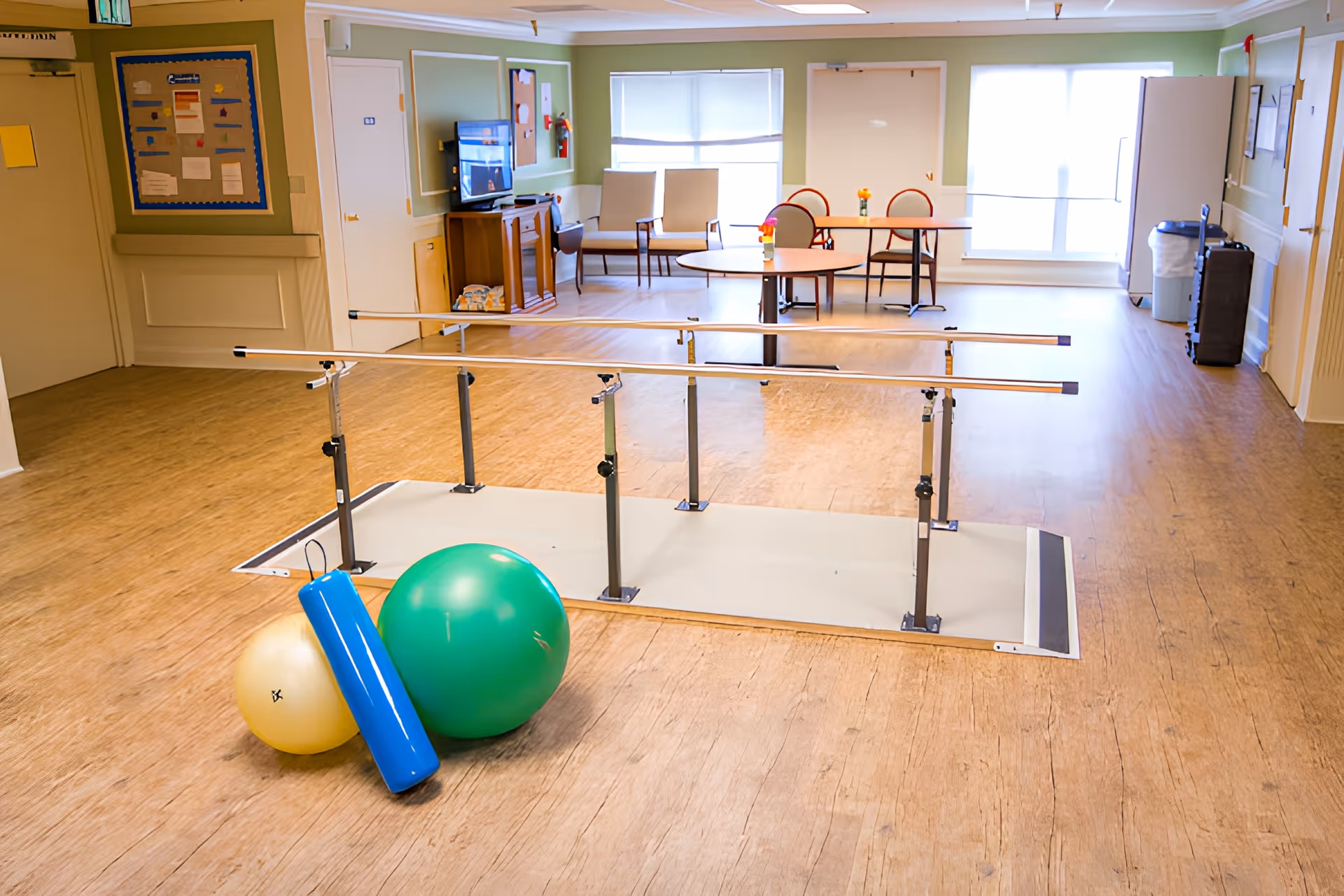 A spacious rehabilitation room with parallel bars for walking exercises, two exercise balls (one green and one yellow), and a blue cylindrical foam roller on a wooden floor. In the background, there are chairs, tables with small flower vases, a television on a wooden stand, and large windows letting in natural light.