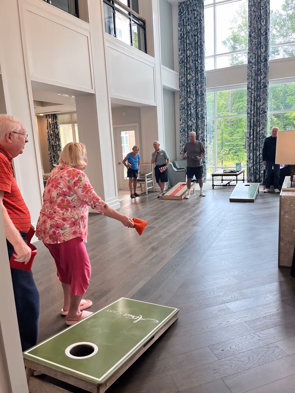 A group of elderly people playing cornhole indoors in a spacious room with large windows and tall floral curtains. The room has wooden flooring and white walls with high ceilings. One woman in a pink floral top and pink pants is throwing a bean bag towards a green cornhole board labeled 'Atria Cary'. Other participants watch and wait their turn.