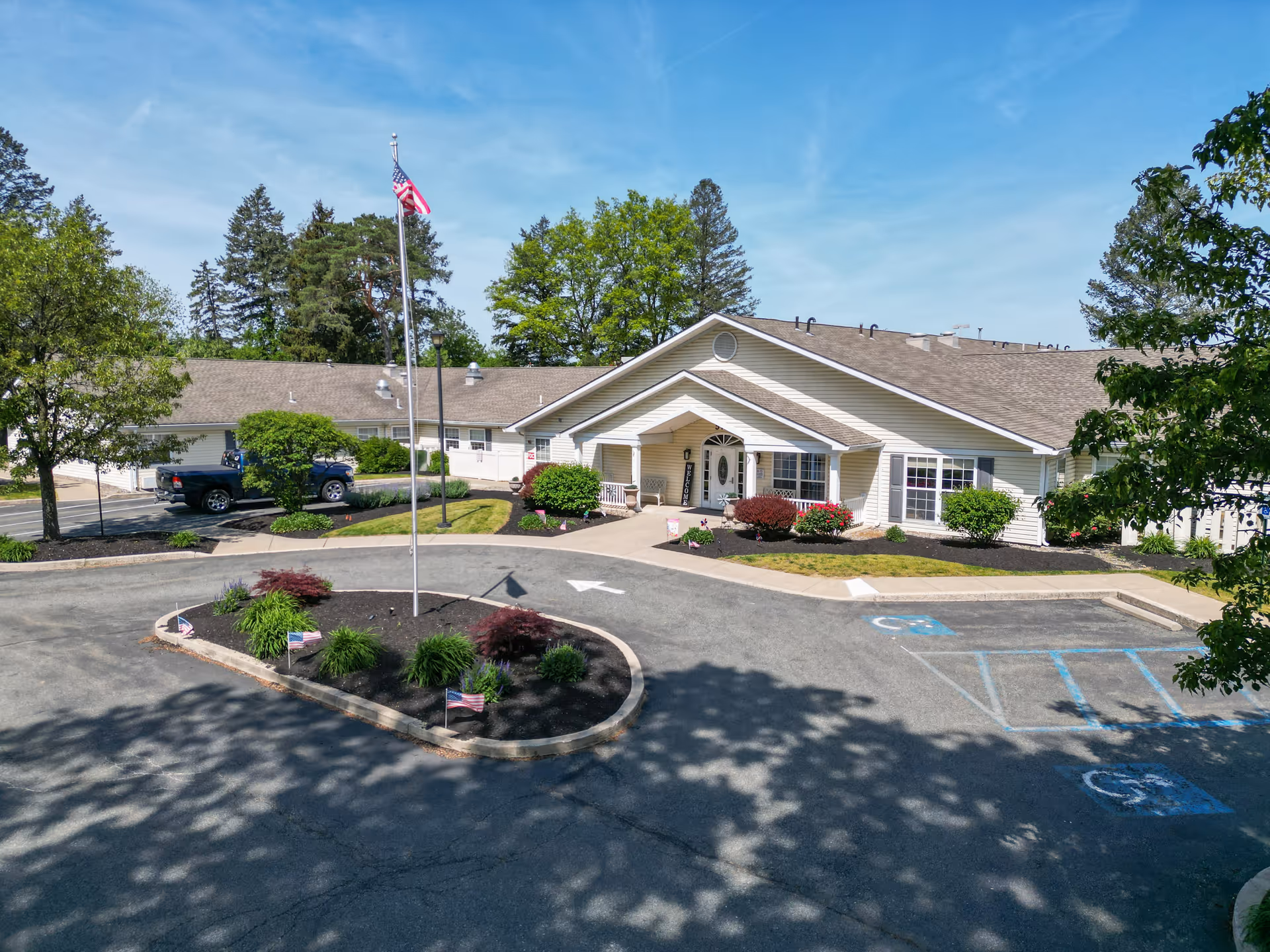 Front exterior view of Arden Courts - ProMedica Memory Care Community in Allentown, showing a single-story building with a peaked roof, an American flag on a flagpole in a landscaped circular driveway, and several bushes and trees around the property under a clear blue sky.
