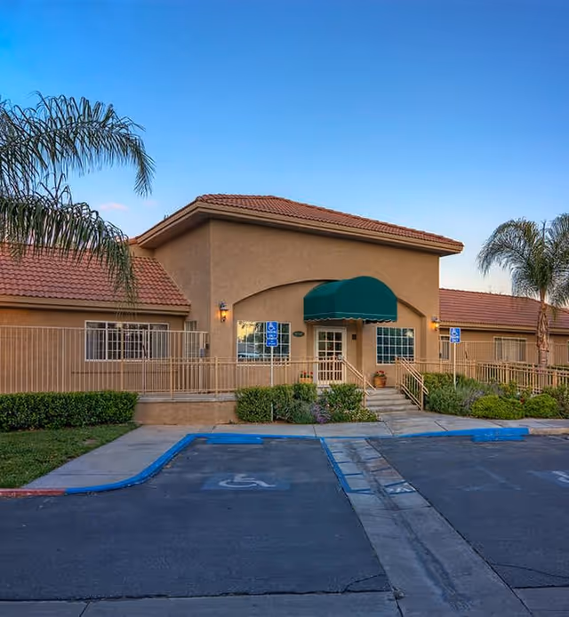 Exterior view of Desert Hills Memory Care Center building with a tan stucco facade, red tile roof, green awning over the entrance, palm trees on either side, and handicap parking spaces in front.