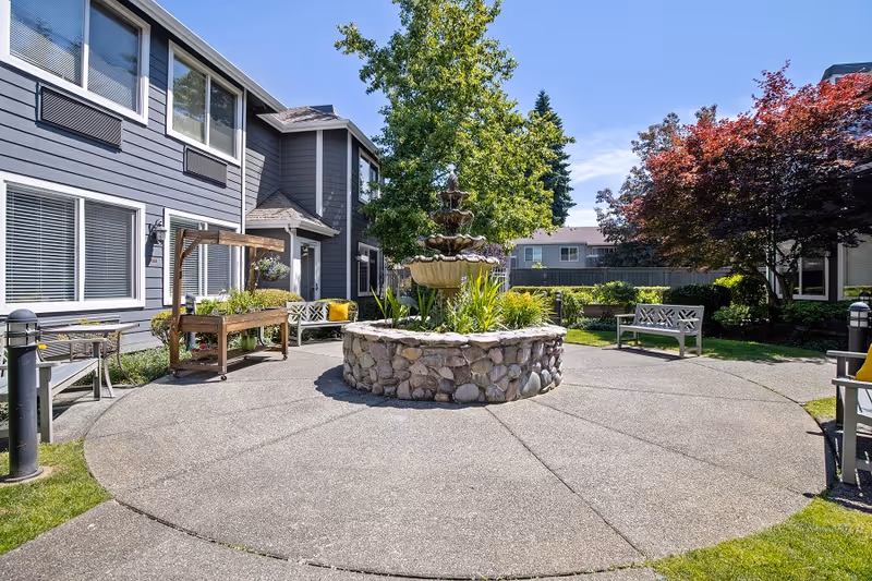 Sunlit courtyard with a central stone fountain, benches, planters, and a gray two-story building in the background.