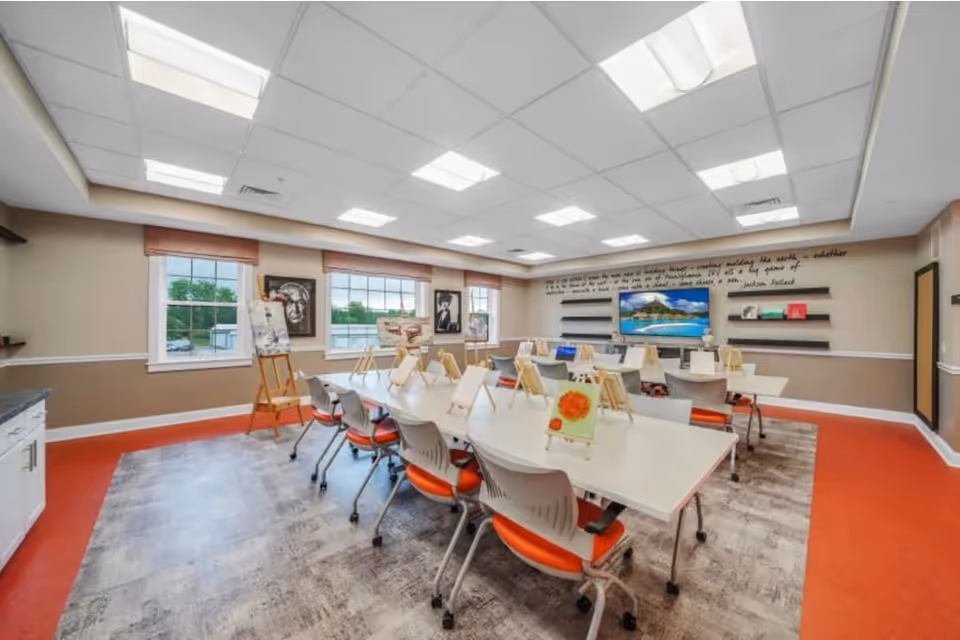 A bright and spacious art activity room with large windows, white tables arranged in a U-shape, and chairs with orange seats. Easels with paintings are placed on the tables and near the windows. The walls have framed artwork and a large TV screen mounted on the far wall, with shelves holding books and decorative items. The room has a mix of carpet and red flooring, and a ceiling with recessed lighting.