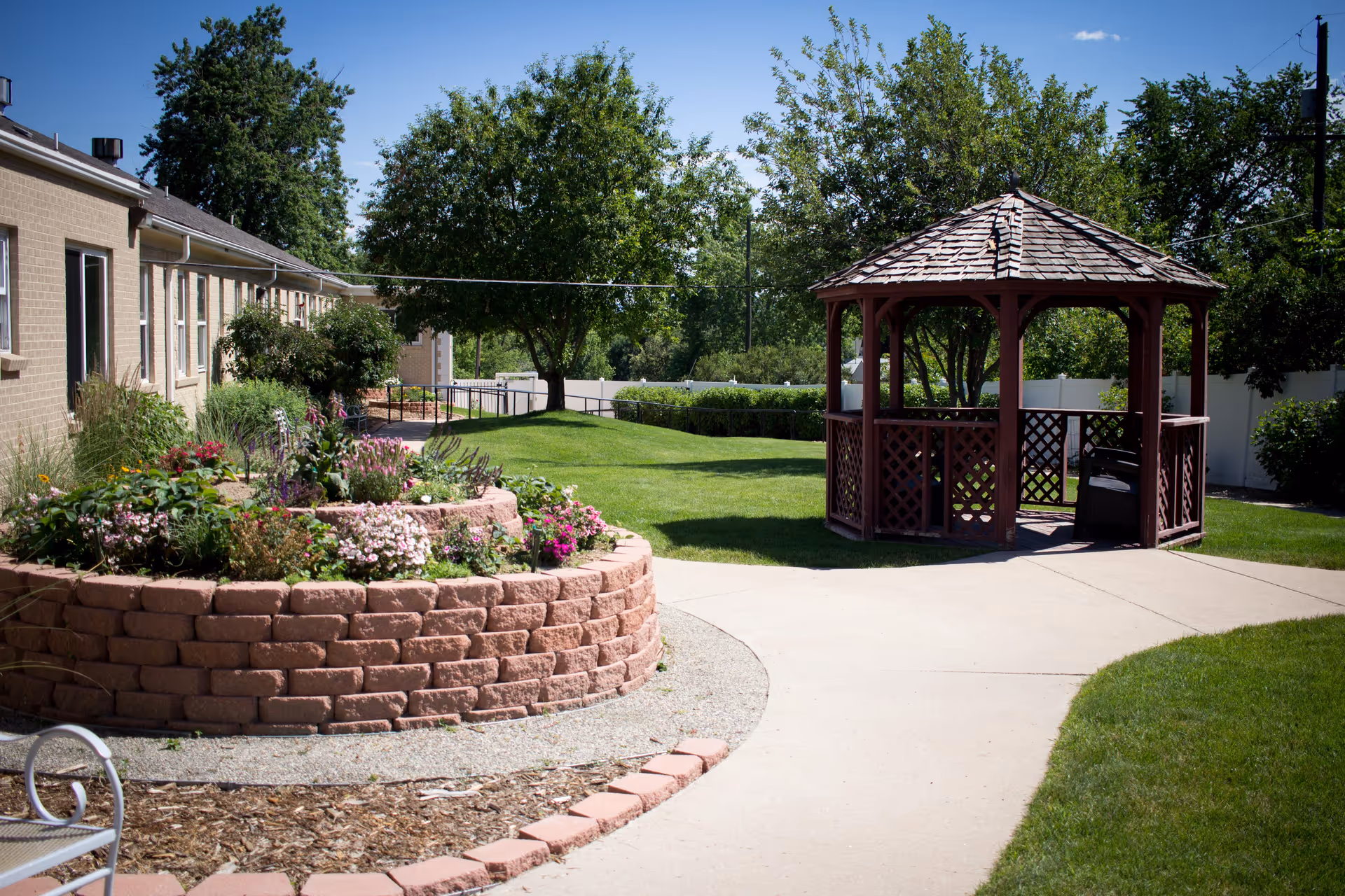 Outdoor garden area at Spring Ridge Park Assisted Living featuring a circular raised flower bed with various colorful flowers, a paved walkway, a wooden gazebo, green grass, and trees under a clear blue sky.