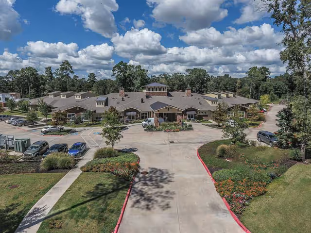 Front exterior view of Bristol Park At Conroe facility showing a large single-story building with a driveway and landscaped garden areas under a partly cloudy blue sky.