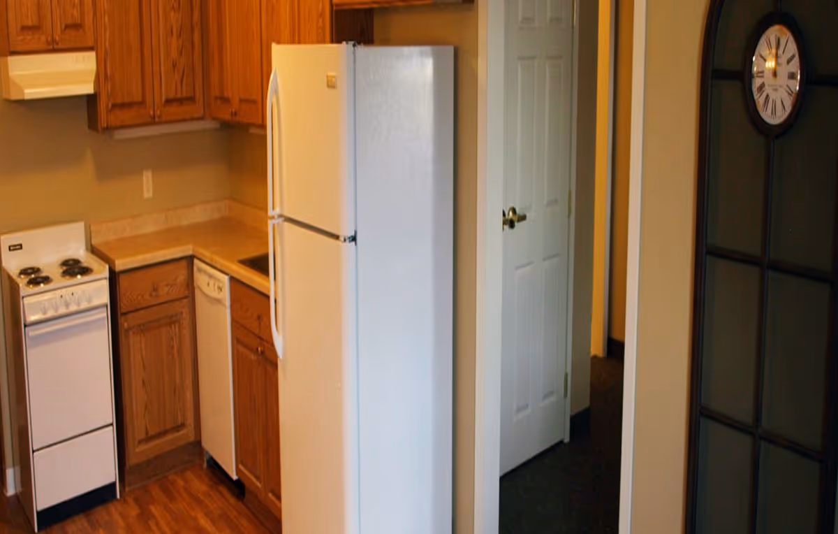 Compact kitchen with a white refrigerator, small electric stove, dishwasher and oak cabinets beside an interior doorway and wall clock.
