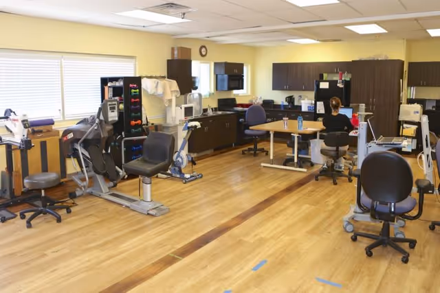 A spacious room with exercise equipment including stationary bikes and weights, a table with chairs, cabinets, and a person sitting at the table facing away from the camera. The room has wooden flooring and yellow walls with windows letting in natural light.