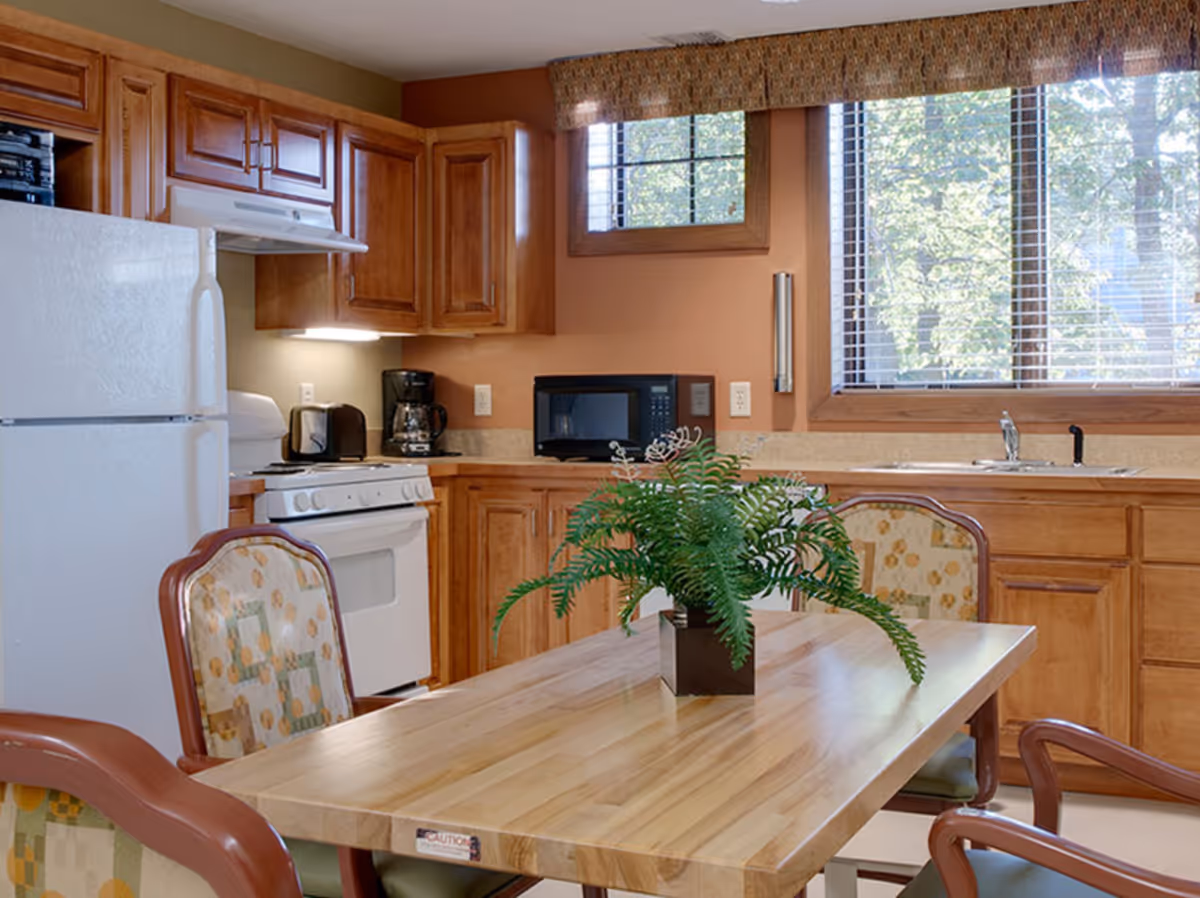 A kitchen interior featuring wooden cabinets, a white refrigerator, a white stove with a range hood, a microwave, a coffee maker, and a toaster. There is a wooden dining table with four cushioned chairs around it, and a green potted plant centerpiece on the table. A large window with blinds allows natural light to brighten the room.