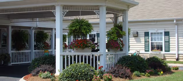 A covered white wooden gazebo/porch with hanging ferns, flower boxes, and manicured shrubs in front of a light-colored building.