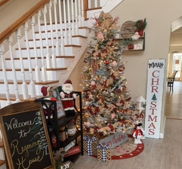 A festive holiday scene inside a senior living facility with a decorated Christmas tree adorned with lights, ribbons, and ornaments. Next to the tree is a small black shelf holding a Santa Claus figure and holiday decorations. A chalkboard sign reads 'Welcome to Regency House.' A tall vertical sign says 'Merry Christmas.' The setting is near a staircase with white railings and wooden steps, and a glimpse of a dining area is visible in the background.