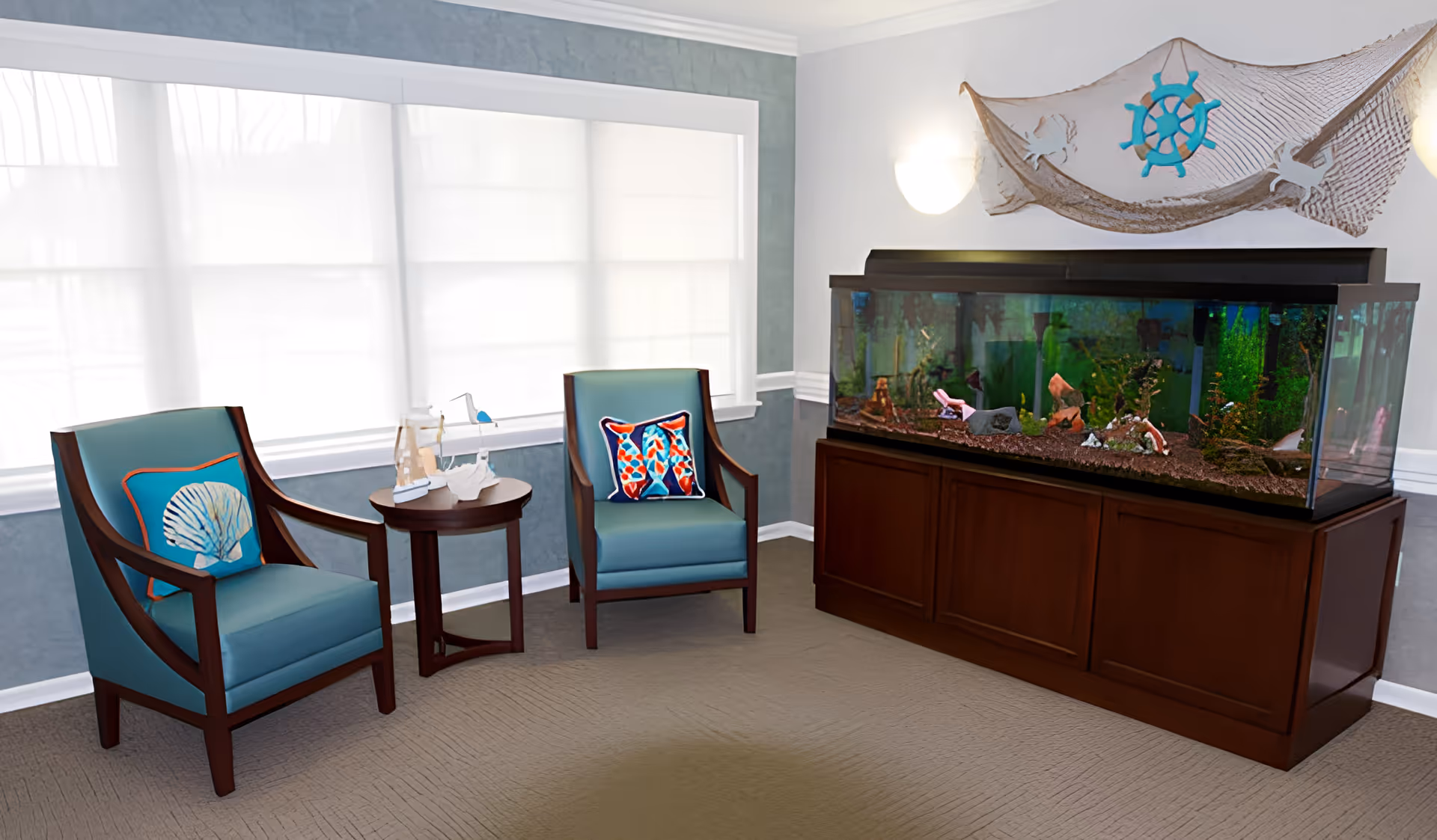 A cozy sitting area with two teal armchairs featuring decorative pillows, a small round wooden table with nautical-themed decor, and a large fish tank on a wooden cabinet. The wall behind the fish tank has a nautical net decoration with a blue ship wheel. The room is softly lit with natural light coming through white window shades.