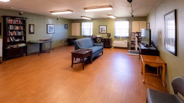 A spacious common room with wooden flooring and green walls, featuring a blue loveseat sofa with a wooden side table, bookshelves filled with books, a dartboard on the wall, a television on a stand, and several windows letting in natural light.