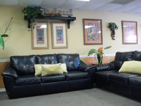 Seating area with black leather sofas, decorative pillows, framed artwork, and potted plants against a beige wall.