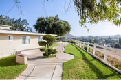 Curved paved walkway alongside a low beige building with manicured shrubs, a bench, white fence, and a distant hillside view.