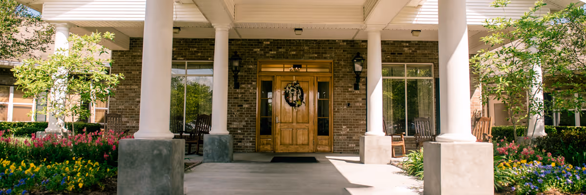 Front entrance of a brick senior living facility with white columns, a wooden double door decorated with a wreath, porch rocking chairs, and flowerbeds.
