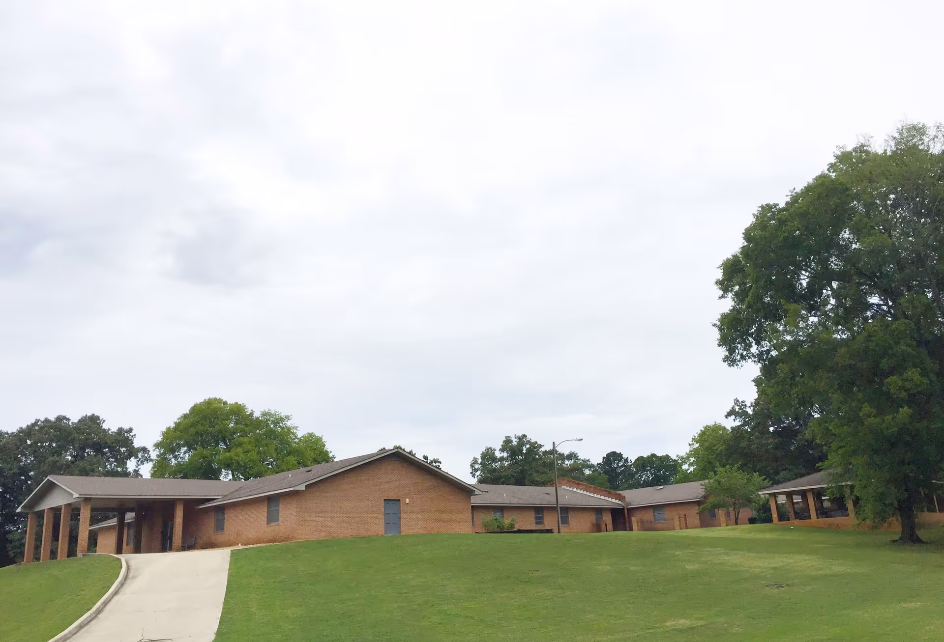 Exterior view of a single-story brick nursing home building with a sloped driveway and green lawn in front, surrounded by trees under a cloudy sky.