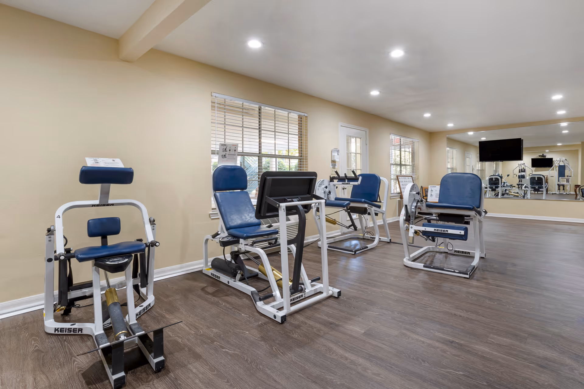 A fitness room with several exercise machines featuring blue padded seats and white frames. The room has wood flooring, beige walls, large windows with blinds, a wall-mounted TV, and a large mirror covering one wall.