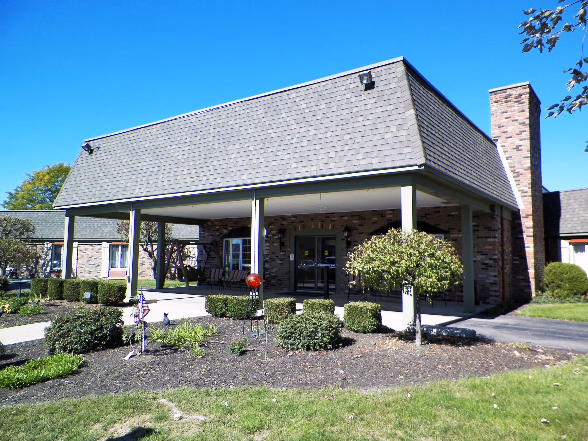 Exterior view of Piqua Manor senior living facility showing a covered entrance with brick walls, a shingled roof, and landscaped bushes and small trees in front under a clear blue sky.
