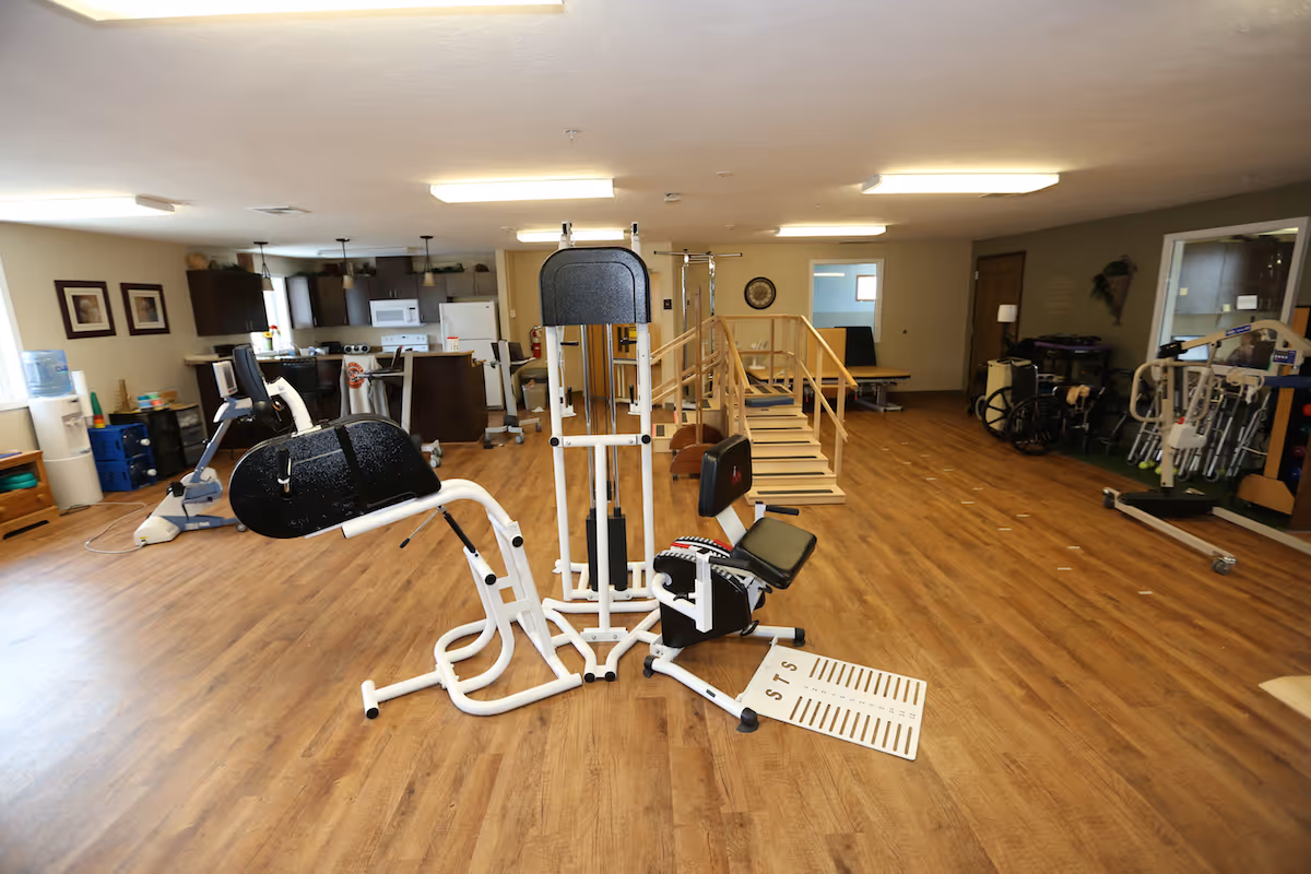 A spacious rehabilitation or physical therapy room with wooden flooring, exercise equipment, a small ramp with handrails, wheelchairs, and walkers. The room has a kitchen area in the background with cabinets, a microwave, and a refrigerator.