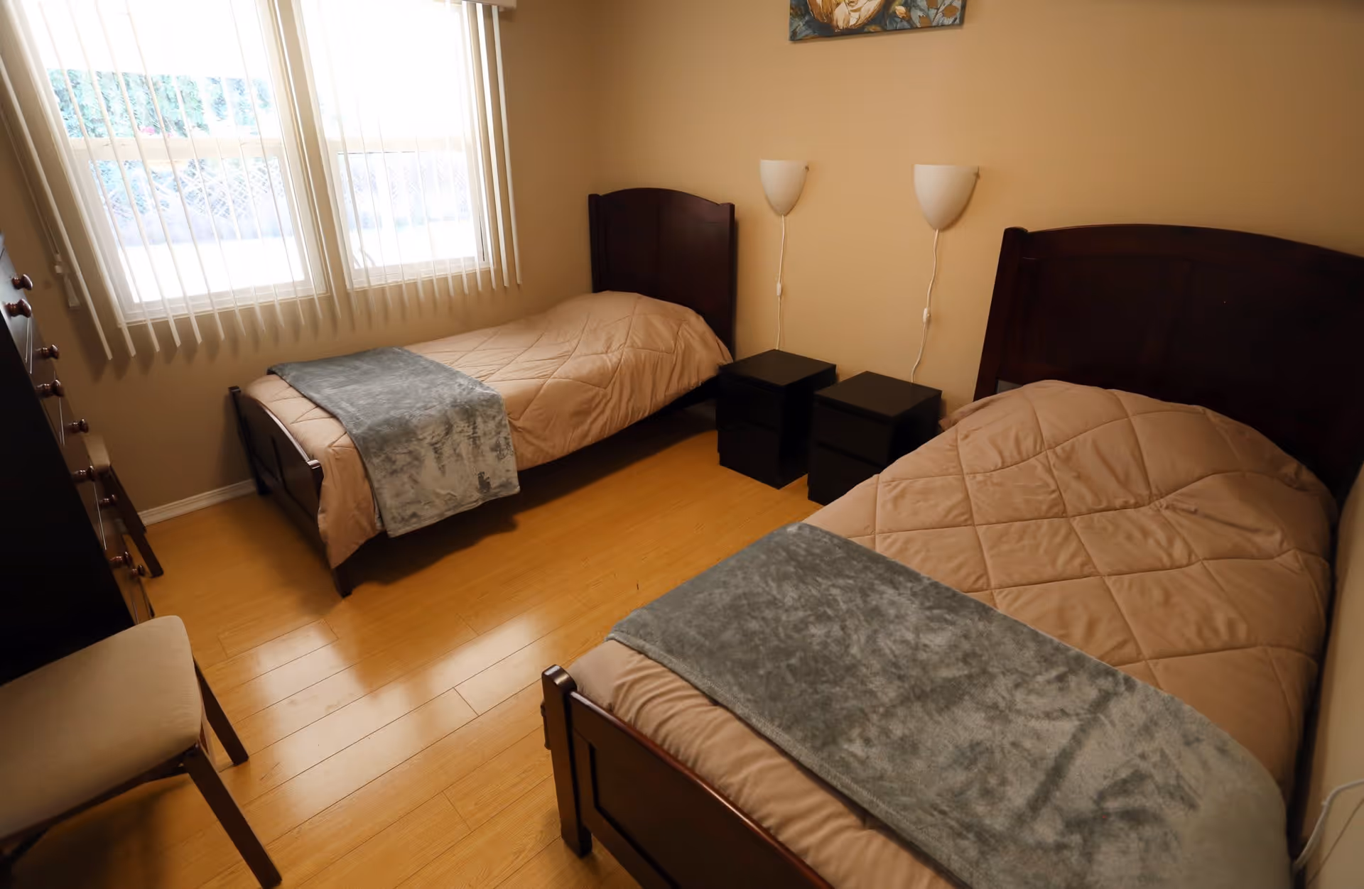 A simple bedroom with two single beds, each covered with beige comforters and gray throws at the foot. Between the beds are two black nightstands with wall-mounted lamps above them. The room has wooden flooring, a window with vertical blinds, a wooden dresser, and a chair.