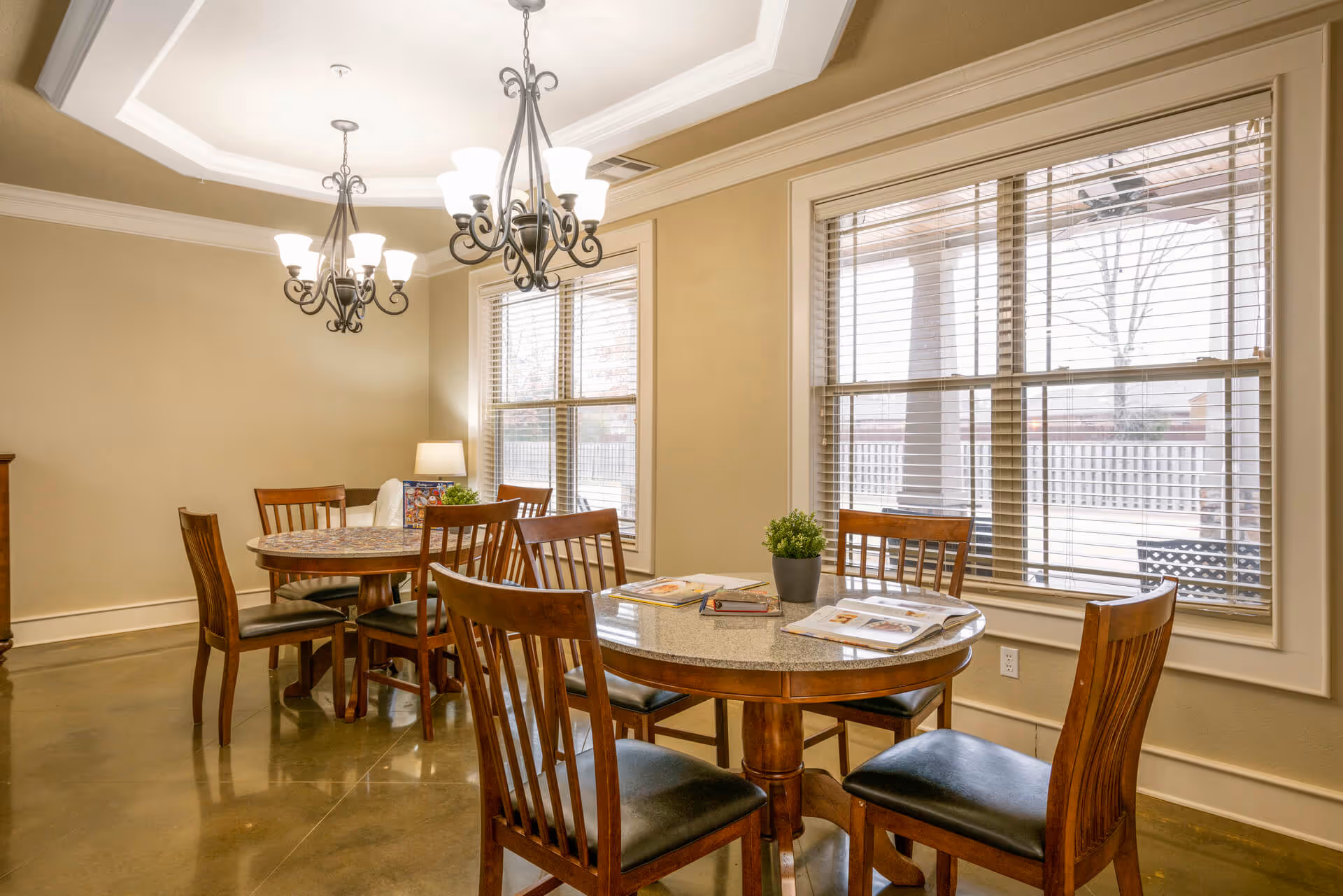 A bright dining room with two wooden dining tables surrounded by wooden chairs with black cushions. The tables have magazines and a small potted plant on them. Large windows with blinds allow natural light to fill the room. Two elegant chandeliers hang from the ceiling, and a lamp is visible in the corner.