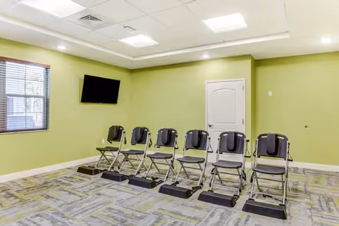 A room with light green walls and a patterned carpet featuring six black folding chairs arranged in a row, each with a black step platform in front. A flat-screen TV is mounted on the wall to the left, and there is a window with blinds and a white door on the far wall. The ceiling has recessed lighting and ventilation panels.