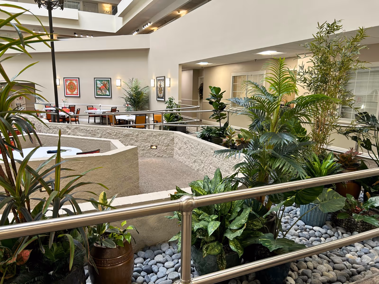 Indoor atrium area of a senior living community featuring a variety of green plants and shrubs in pots surrounded by decorative stones. There is a metal railing in the foreground and a seating area with tables and chairs in the background. The walls are light-colored with framed artwork and wall-mounted lights, and windows overlook the space.