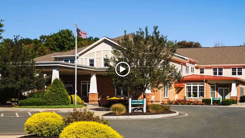 Front exterior of a two-story senior living building with an American flag, circular driveway, and landscaped shrubs.