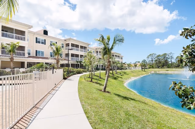 A bright outdoor scene showing a paved walkway lined with palm trees and a white fence on the left, leading to a multi-story residential building with balconies. On the right side, there is a grassy area sloping down to a pond with a water fountain, under a partly cloudy blue sky.