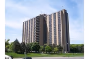 A tall multi-story residential building with numerous windows, surrounded by green trees and a grassy area under a partly cloudy sky.