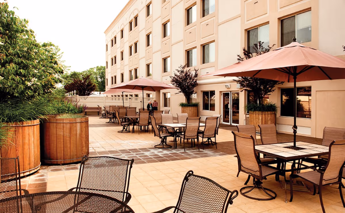 Outdoor patio area with multiple tables, chairs, umbrellas, and large planters beside a multi-story building.
