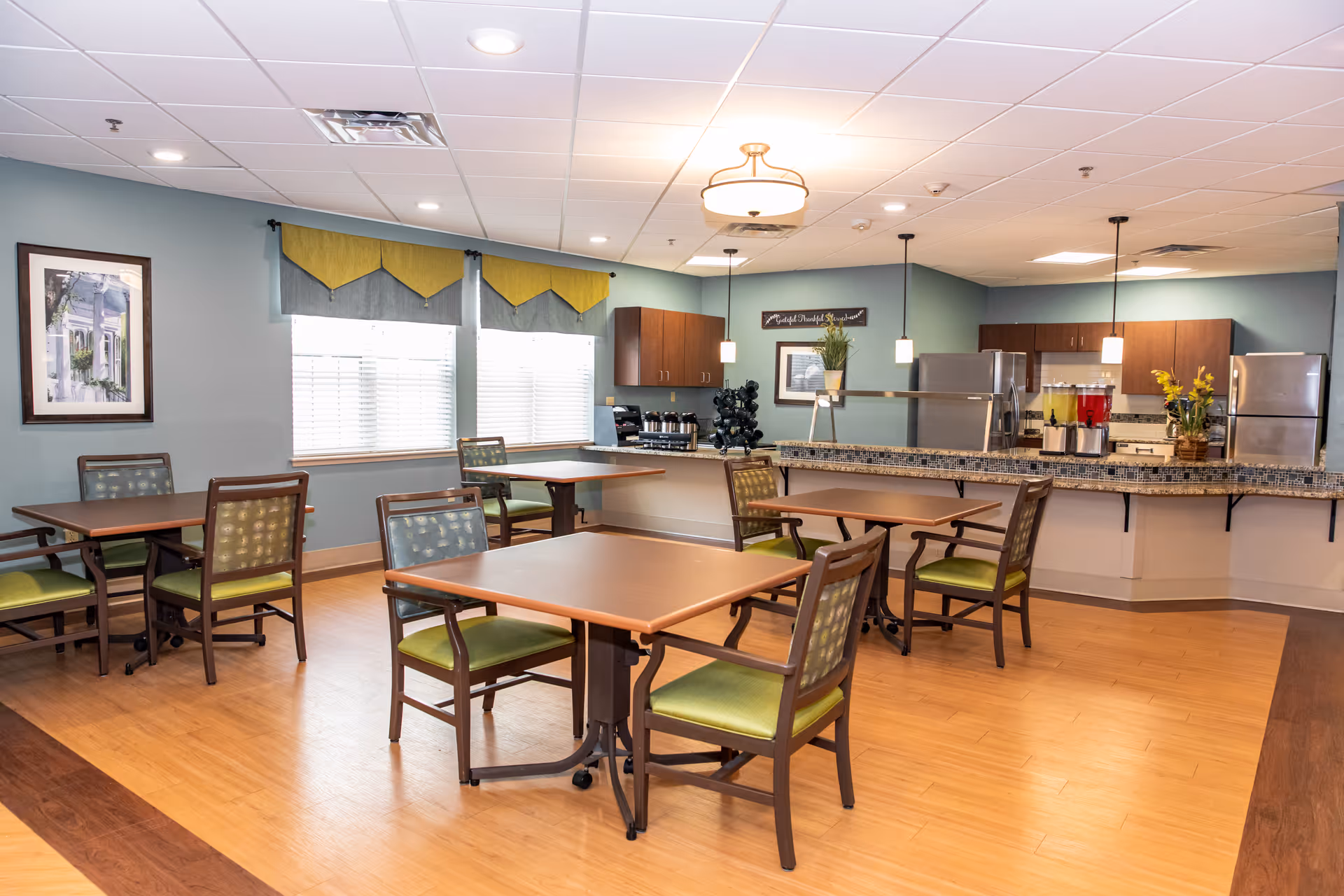 A dining area in a senior living facility with several square wooden tables and green cushioned chairs. The room has light blue walls, large windows with yellow and gray valances, and a kitchen area in the background with stainless steel appliances, a granite countertop, and beverage dispensers. The ceiling has recessed lighting and hanging pendant lights.