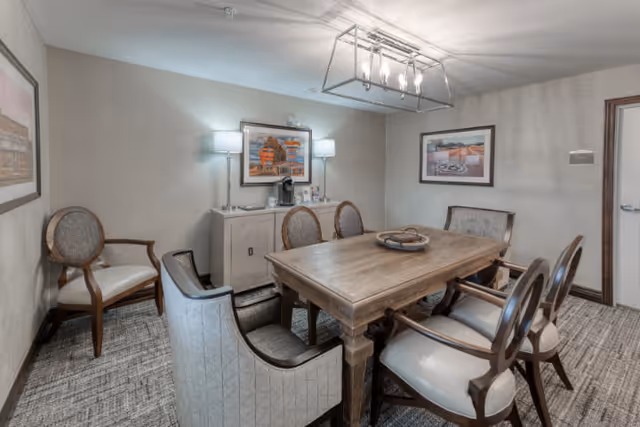 A cozy dining room with a wooden rectangular table surrounded by six cushioned chairs. The room has neutral-colored walls adorned with framed artwork. Two wall-mounted lamps flank a small cabinet with a coffee maker on top. A modern rectangular chandelier hangs from the ceiling, illuminating the space.