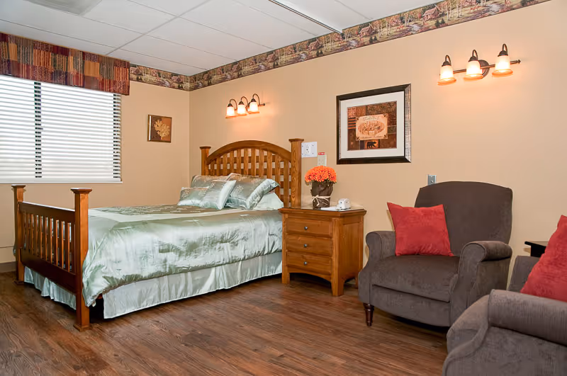 A cozy furnished bedroom in a senior living facility with a wooden bed, nightstand, two armchairs, and wall-mounted lights.